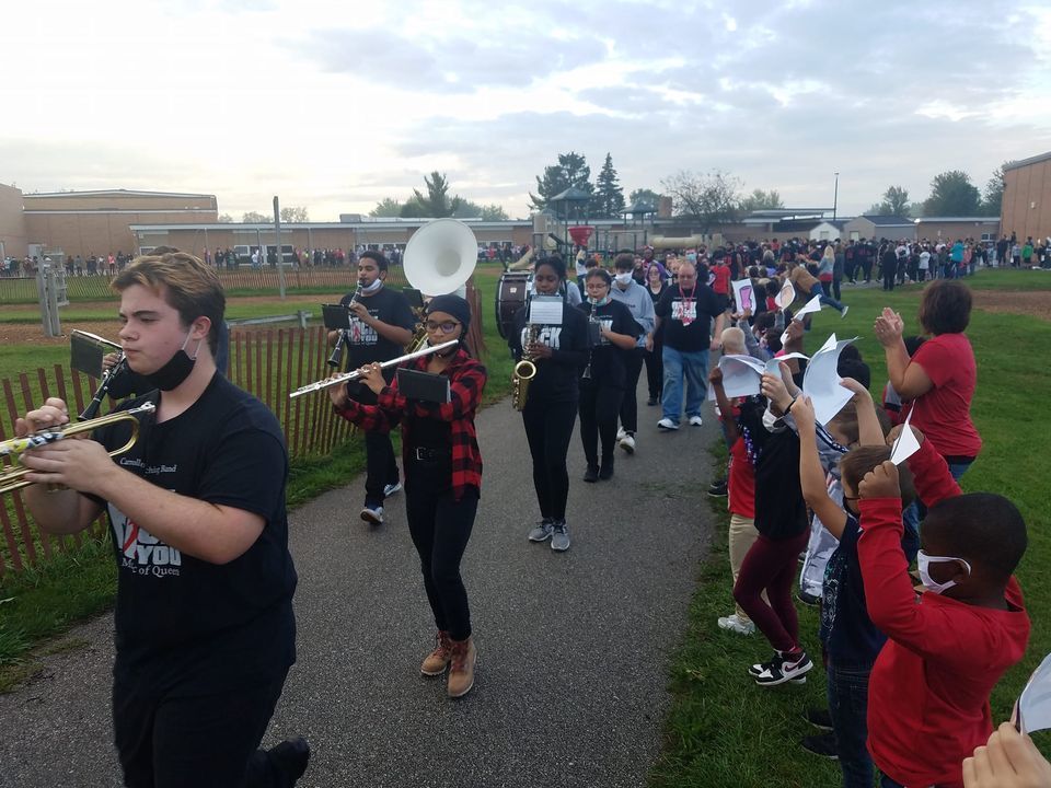 Students marching and playing instruments while younger children hold signs on the sideline